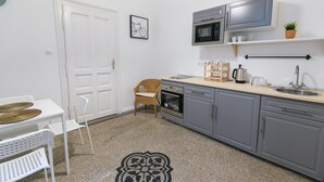 Une cuisine compacte avec des meubles gris, un four à micro-ondes et un évier. On y trouve une chaise en osier et une table blanche avec des sets de table en fibres tressées.