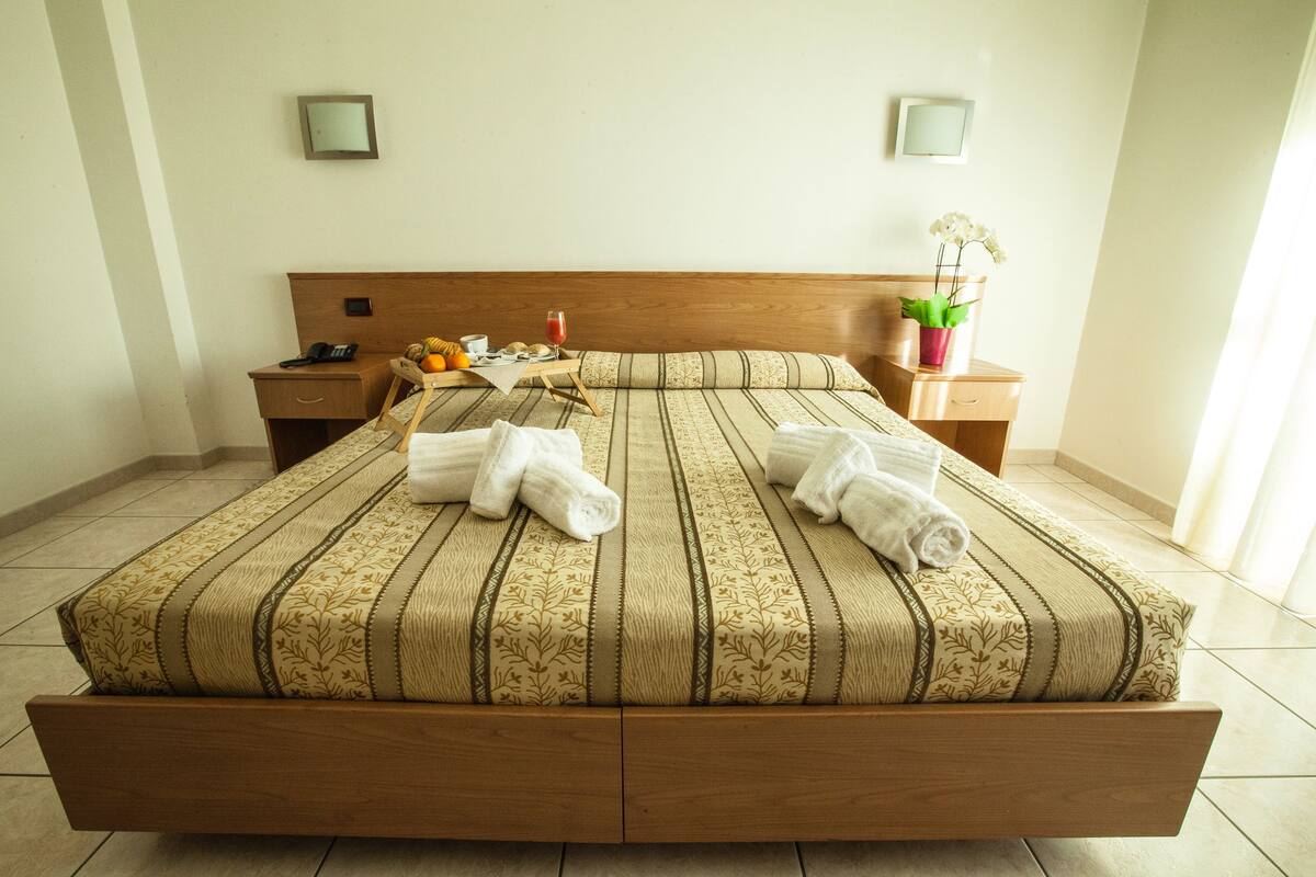 A neatly made bed with a patterned bedspread and two rolled towels, a bedside table with a phone and a small vase of flowers, and a nightstand with a lamp.