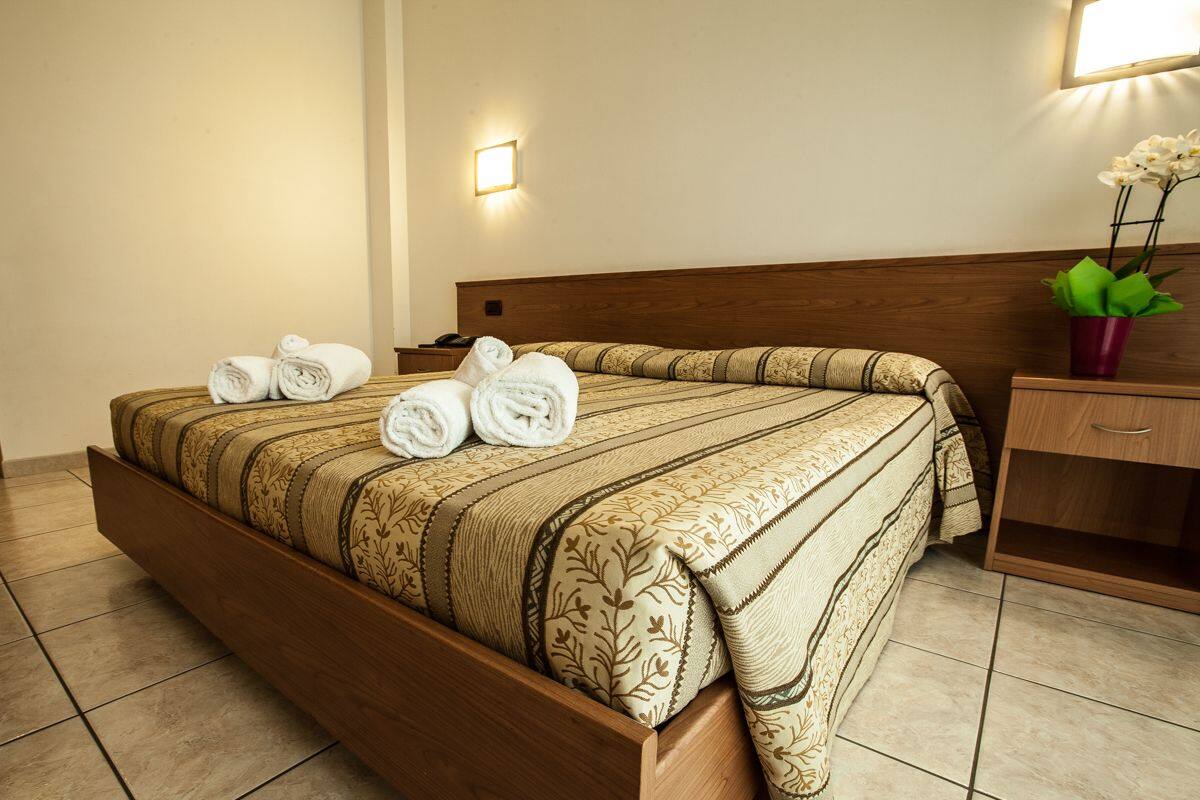 A neatly made bed with rolled towels, a wooden headboard, a bedside table with a lamp, and a potted plant.