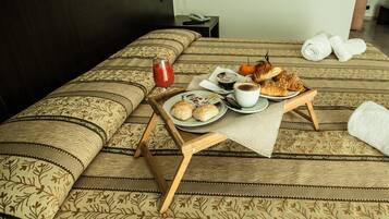 Una cama cuidadosamente tendida con una colcha de motivos florales, una bandeja con artículos de desayuno, incluyendo croissants, un vaso de jugo rojo y una taza de café.