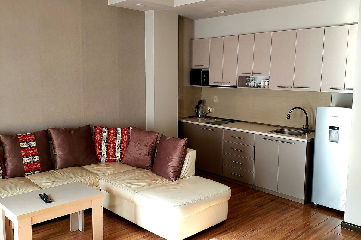 A compact living space with a sofa, ottoman, and a small table. A kitchenette is visible with a refrigerator, microwave, and sink.