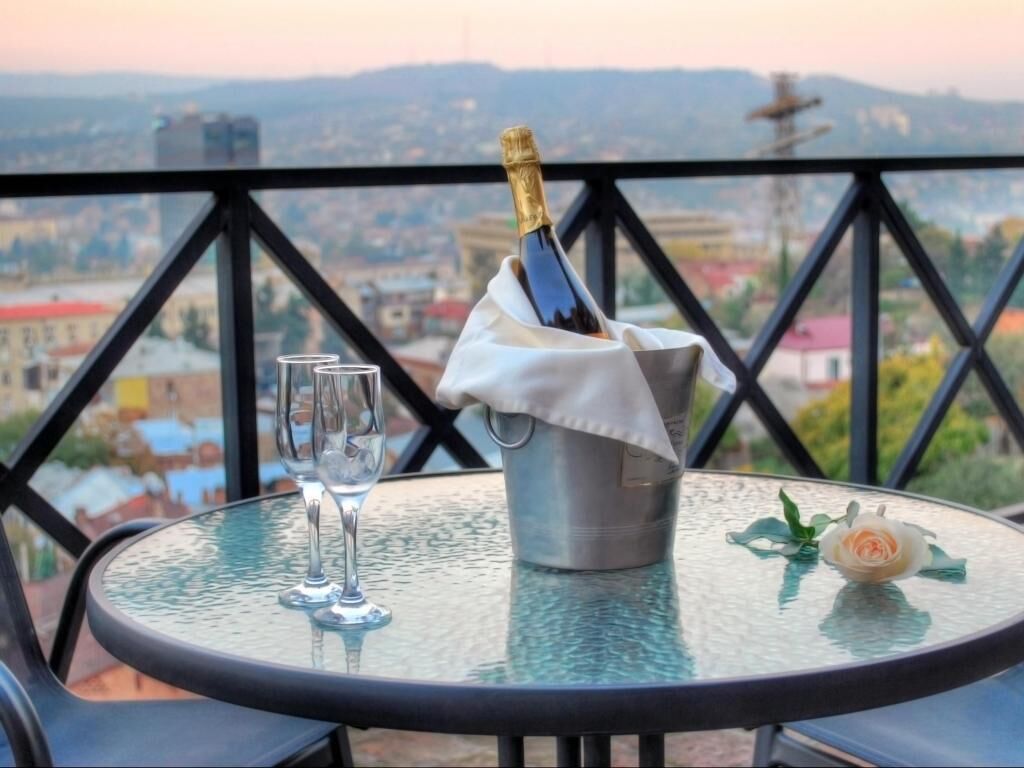 A table with a bottle of champagne, two glasses, and a rose on a balcony overlooking a cityscape.