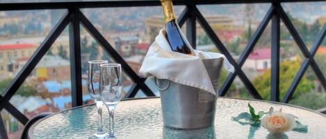 A table with a bottle of champagne, two glasses, and a rose on a balcony overlooking a cityscape.