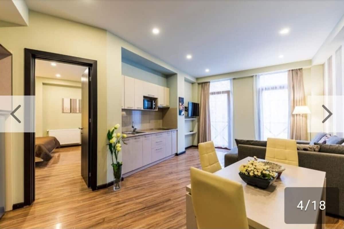 A modern hotel room with a wooden floor, a dining area with a table and chairs, a sofa, a kitchen area, and a view of the city.