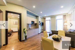 A modern hotel room with a wooden floor, a dining area with a table and chairs, a sofa, a kitchen area, and a view of the city.
