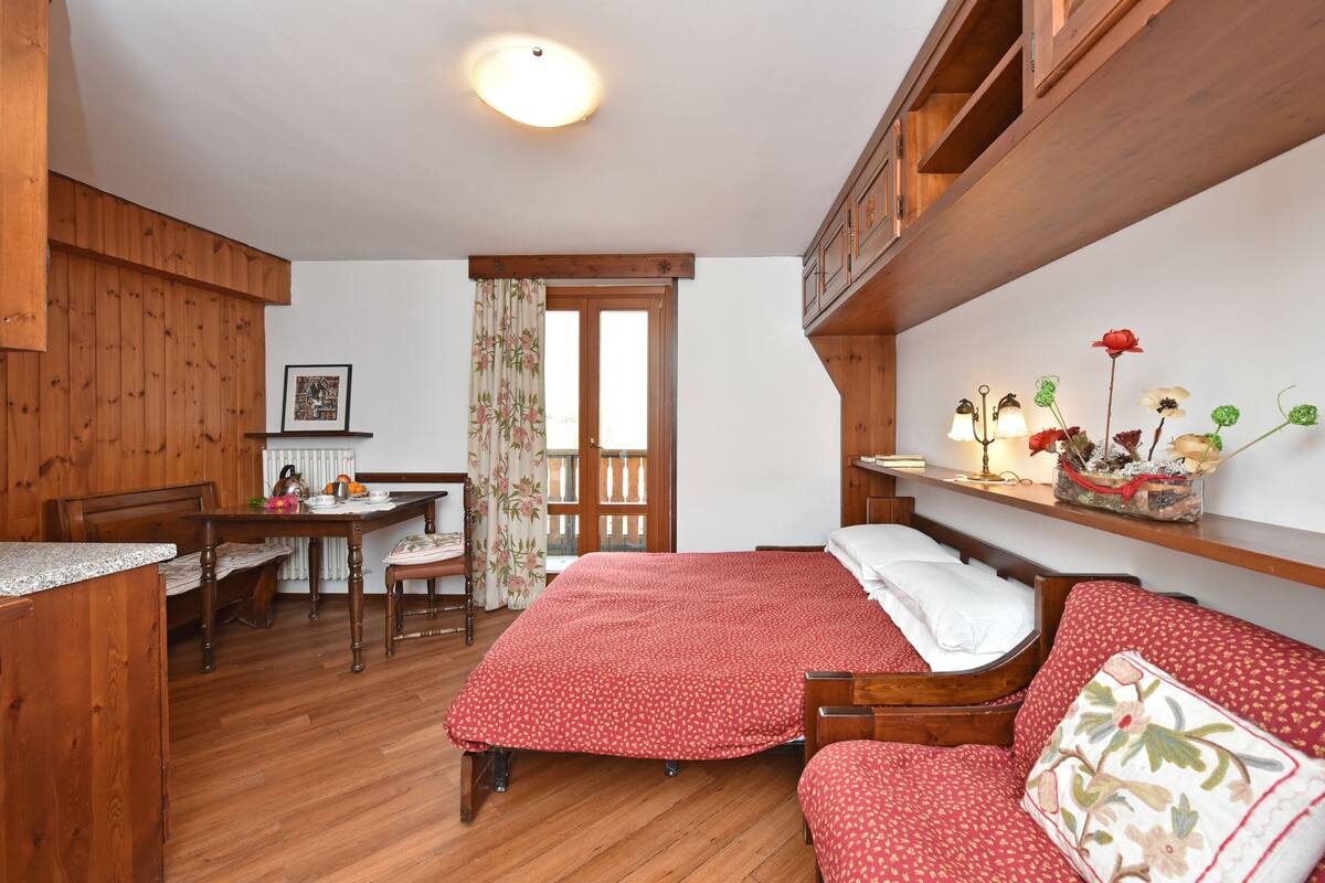 Una habitación con piso de madera, una cama, un banco de madera, una mesa y un ventanal con cortinas.