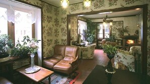 A cozy living room with a leather sofa, a wooden coffee table, and floral wallpaper. There is a fireplace in the background and a large window with lace curtains. - Residence Castelli (Valtournenche)