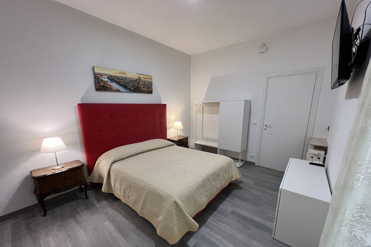 A hotel room with a bed, a red headboard, a nightstand with a lamp, a wardrobe, and a television mounted on the wall.