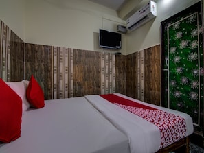 A hotel room with a bed, red pillows, a TV, an air conditioner, and a green door with a floral pattern.