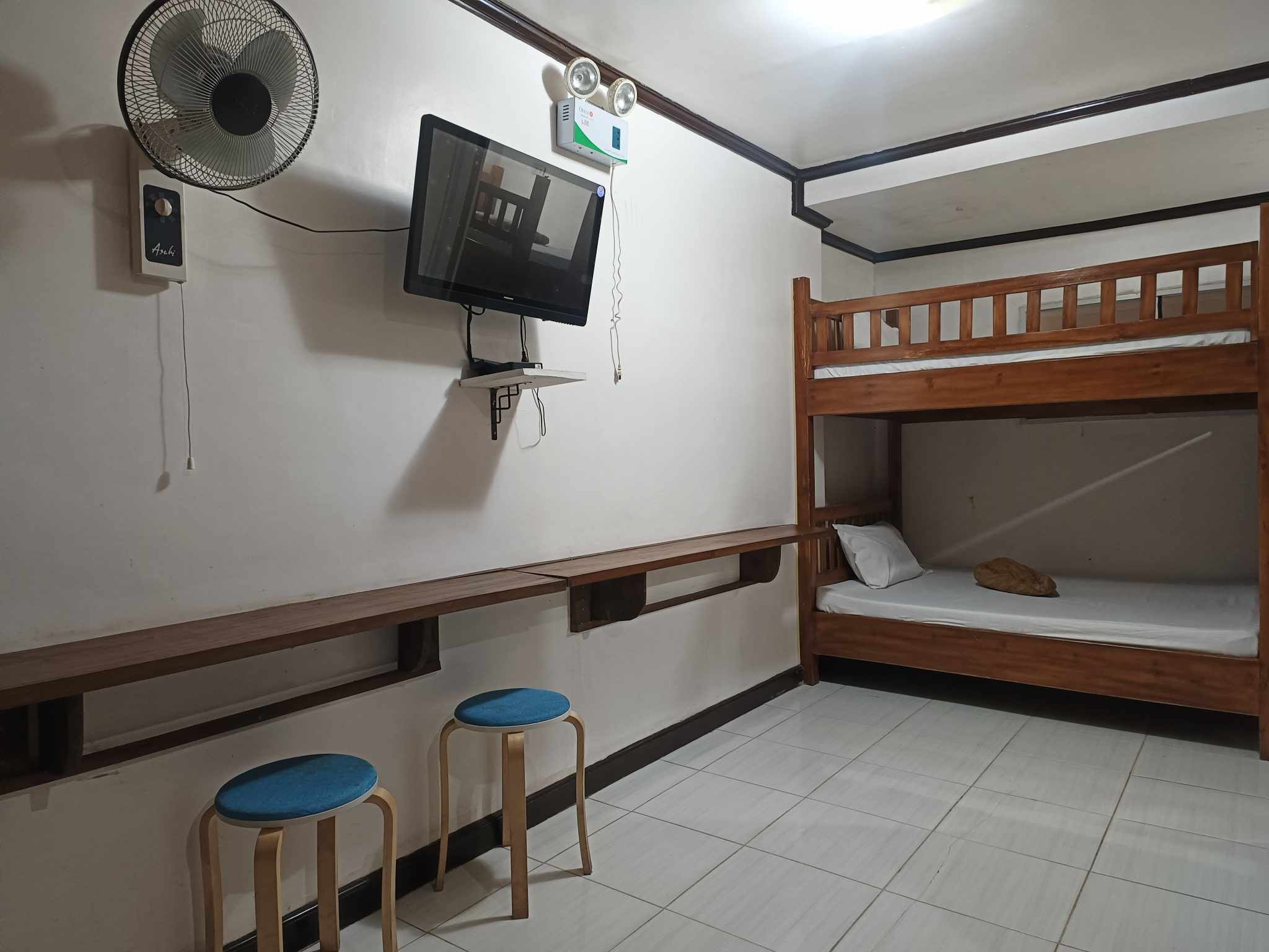 Una habitación con literas, televisión, ventilador y dos taburetes.