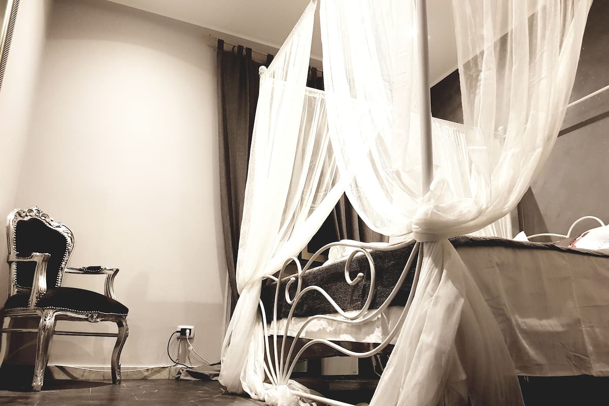 A room with a bed covered by white curtains, a vintage chair, and a bedside table with a lamp.