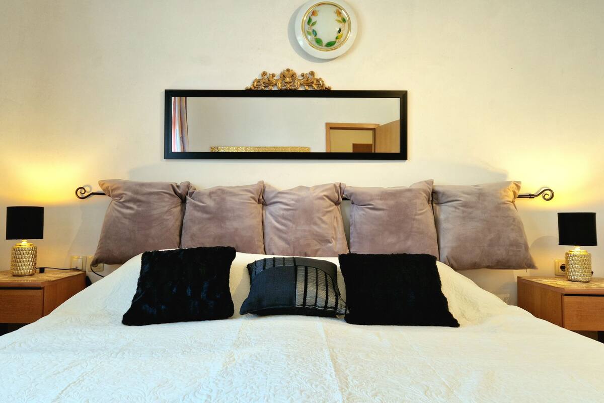 A neatly made bed with a white duvet and multiple pillows, flanked by two bedside tables with lamps, a mirror above the bed, and a decorative plate on the wall.