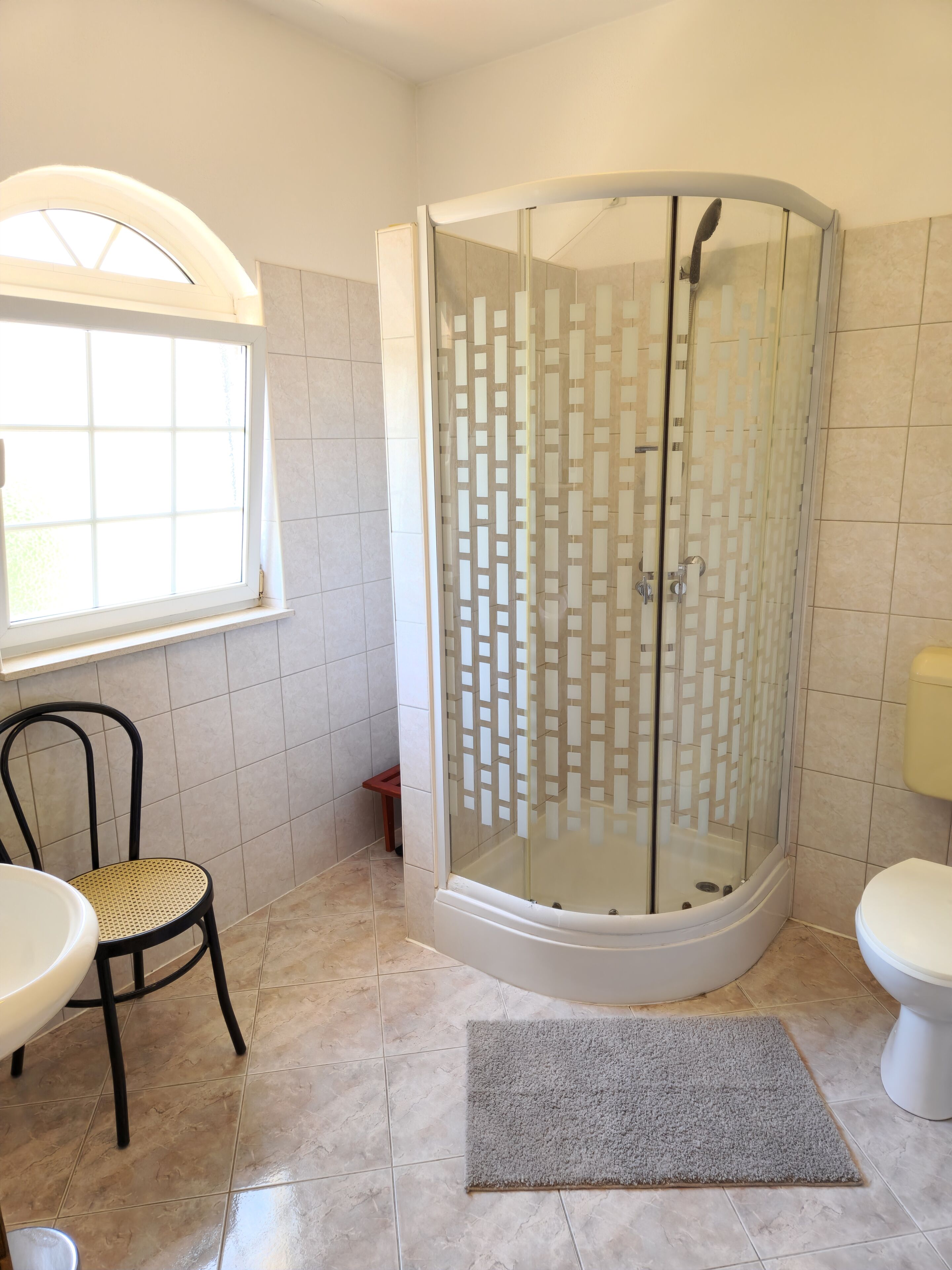 Un baño con ducha de vidrio, inodoro, lavamanos, una silla y una ventana.