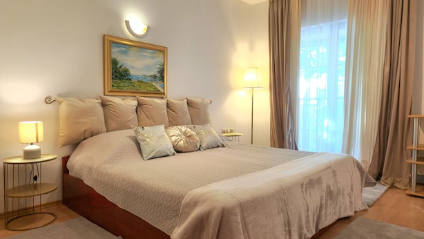 A neatly arranged bedroom with a large bed, two bedside tables, a wall-mounted lamp, and a painting above the bed. - Avalena Rooms (Zadar)