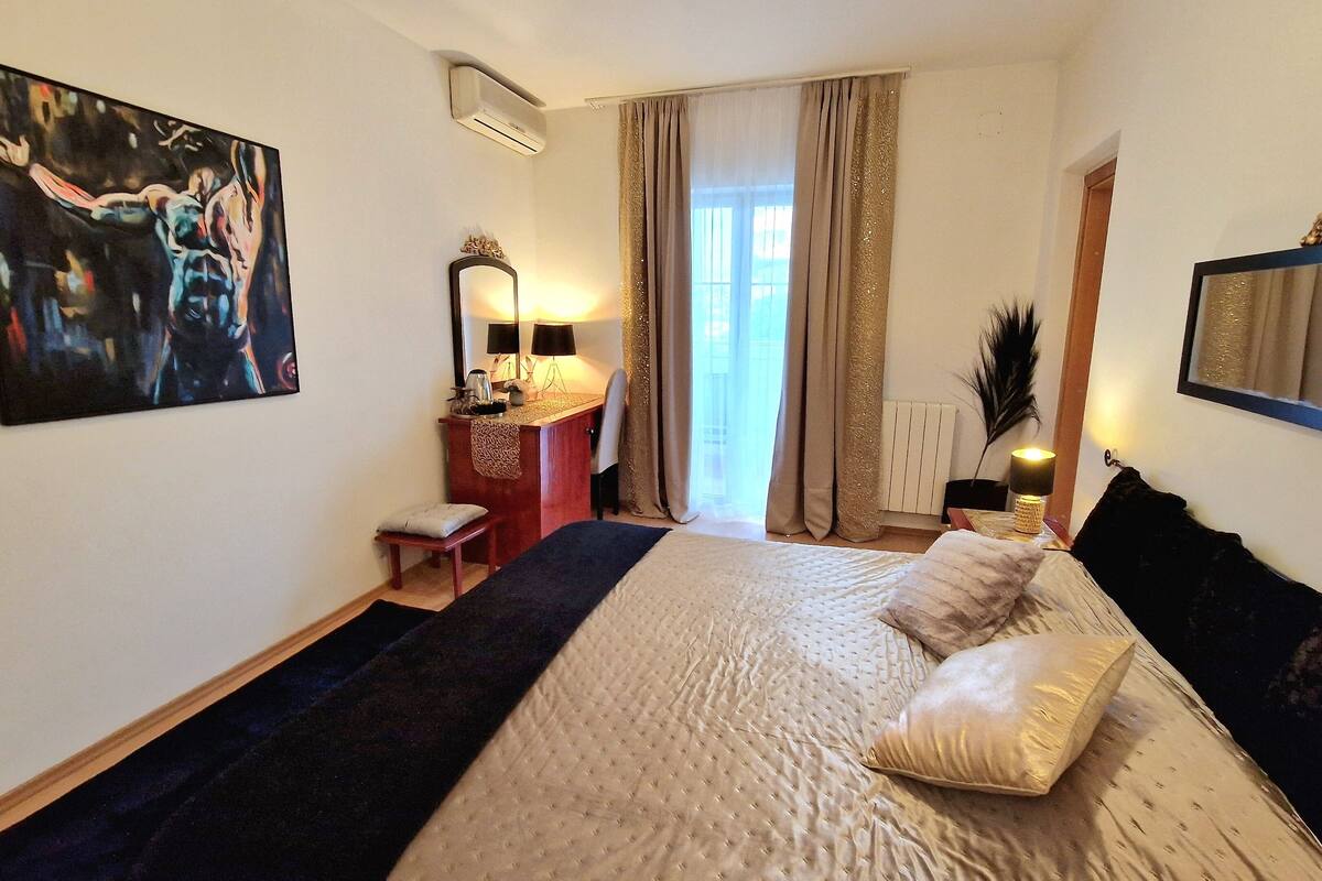 A hotel room with a large bed, a desk with a lamp, a chair, a window with curtains, and a painting on the wall.