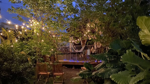 An outdoor seating area with wooden chairs and tables, surrounded by lush greenery and string lights. - Okinawa starry forest cottage (Onna)