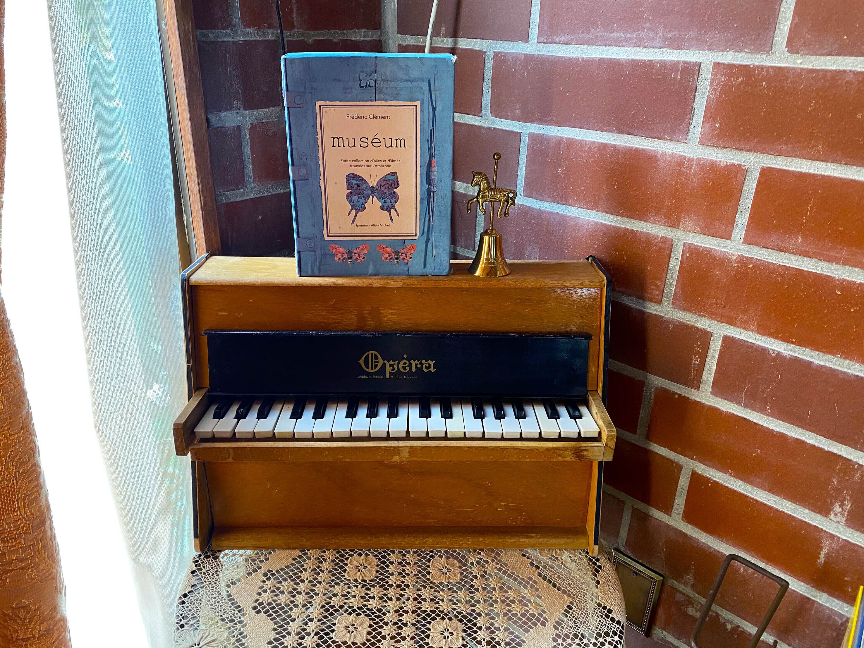 A vintage upright piano with a book titled 'museum' and a golden bell on top, placed against a brick wall.