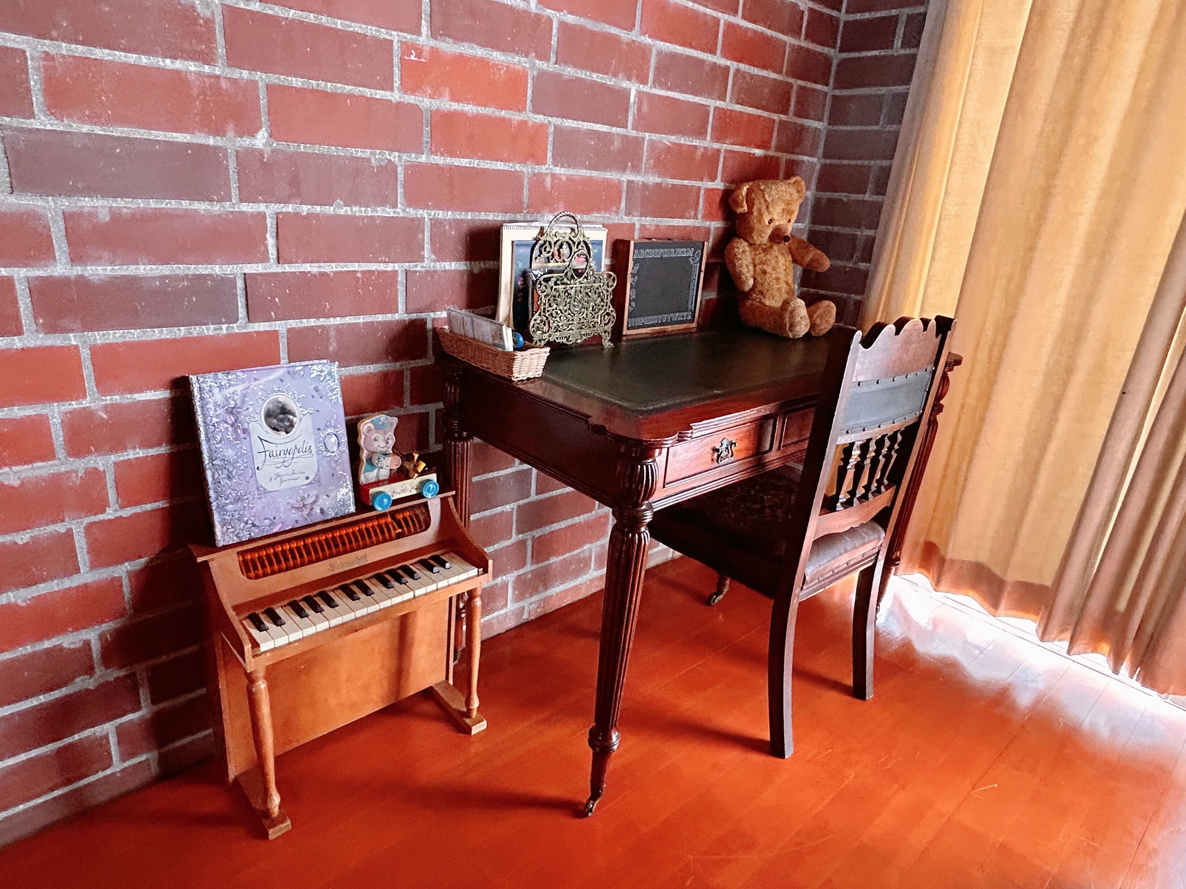 A room with a brick wall, a wooden desk, a chair, a small piano, and a teddy bear.