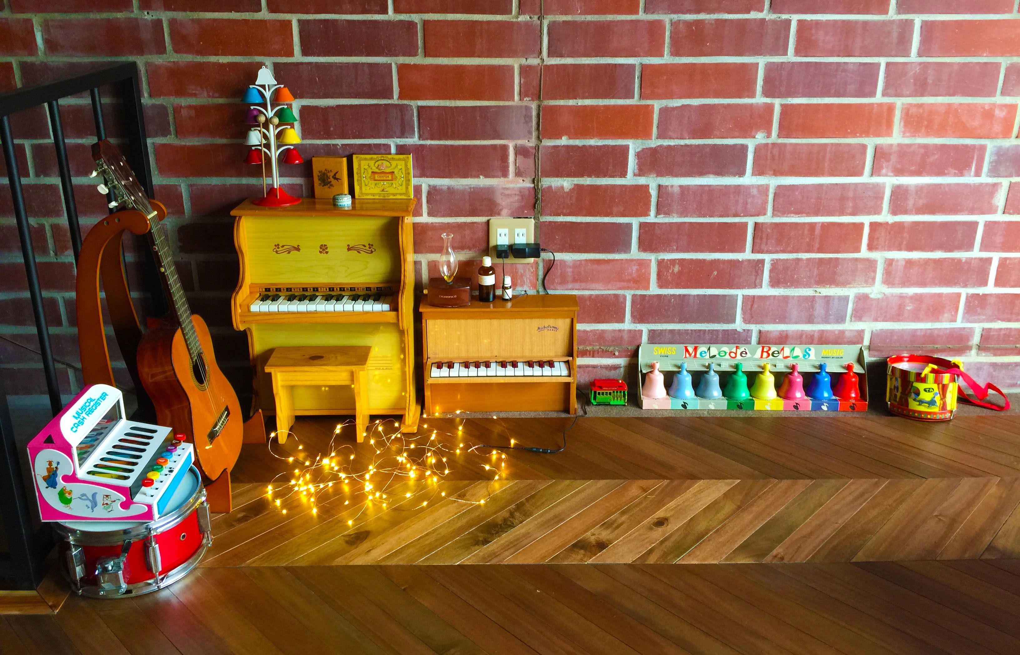 A room with a brick wall, a wooden floor, a piano, a guitar, a toy piano, and a toy drum set.