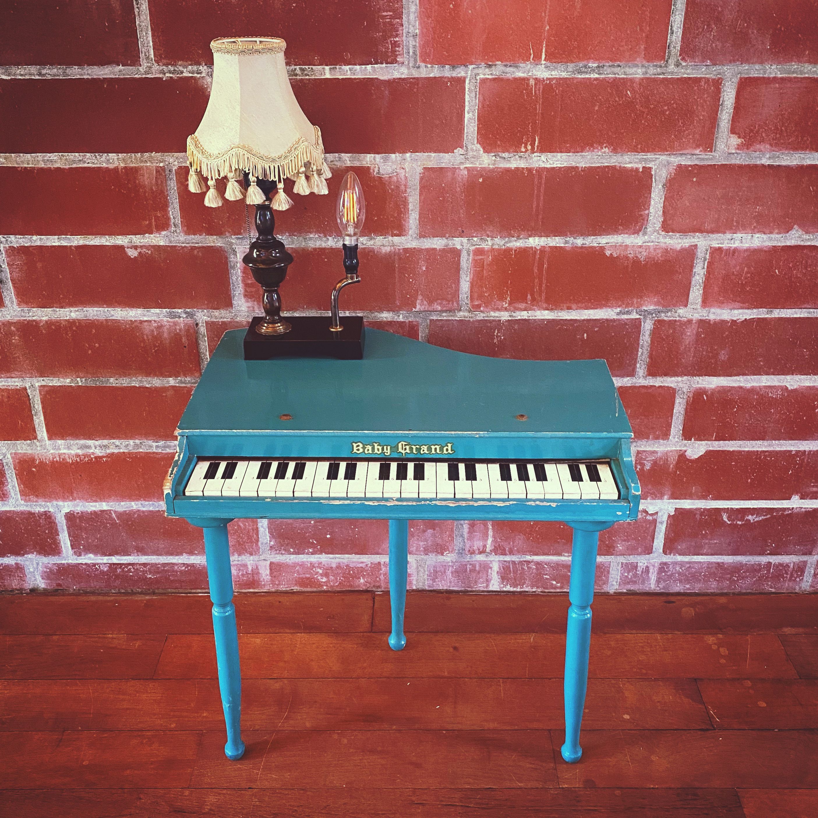 Un piano à queue bleu, de taille réduite, avec une lampe posée sur le dessus, placé contre un mur de briques.