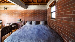 A bedroom with a brick wall, wooden ceiling beams, a bed with a patterned bedspread, a small table with a lamp, and a window. - Okinawa starry forest cottage (Onna)