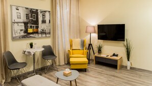 Habitación de hotel con un sillón amarillo, dos sillas negras, una mesita redonda con una caja de pañuelos, una lámpara, un televisor y un cuadro en la pared.