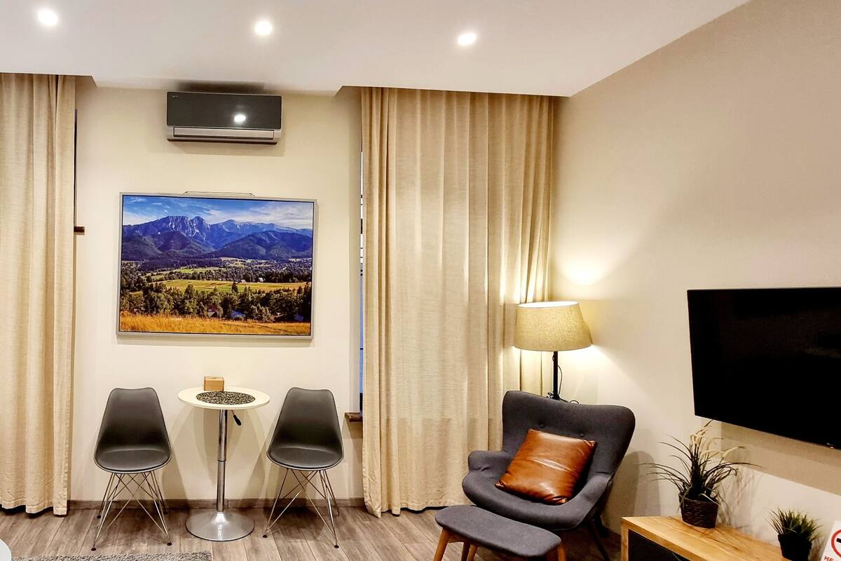 Habitación de hotel con cama, dos sillas, una mesita, televisor y ventana grande con cortinas.