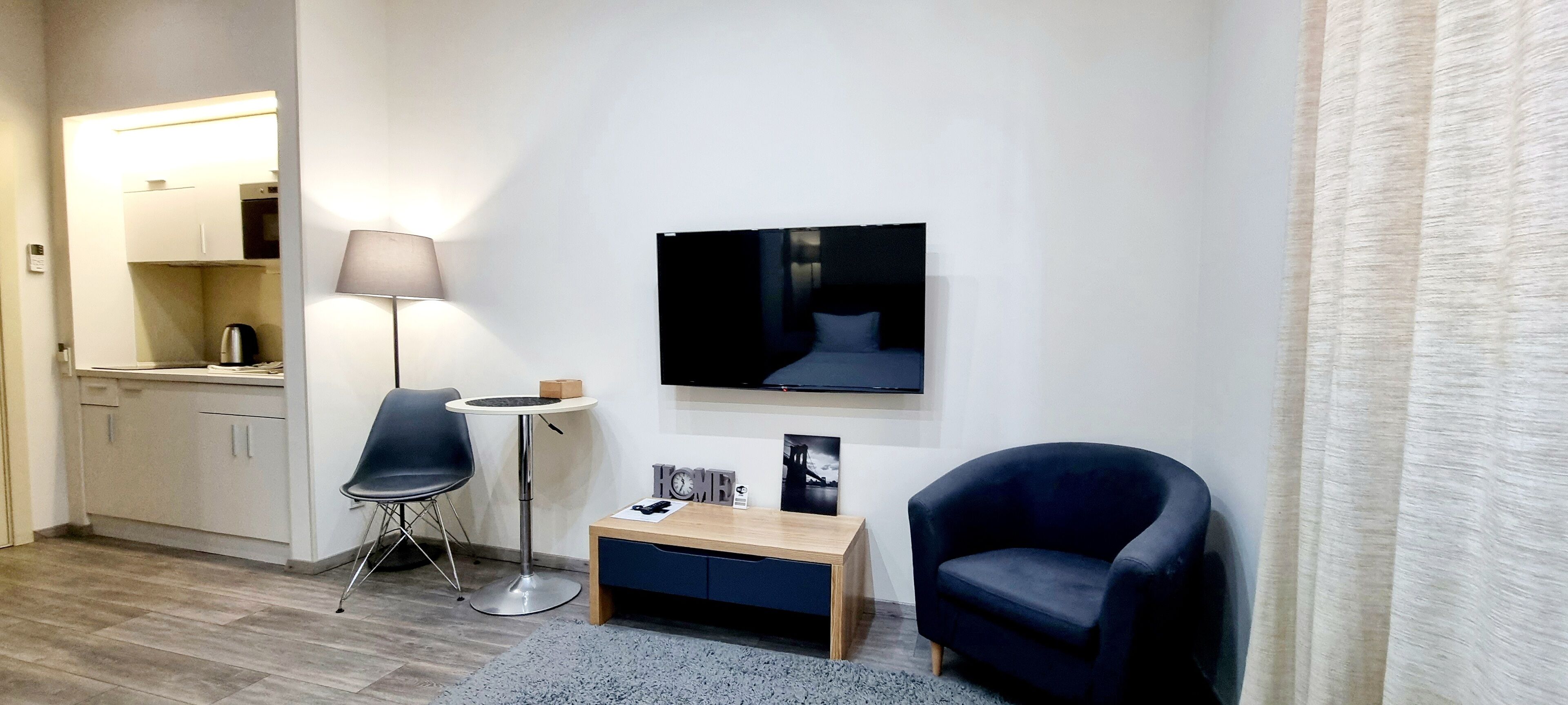 Una habitación de hotel moderna con kitchenette, un televisor de pantalla plana, un escritorio con lámpara, una silla y una alfombra pequeña.