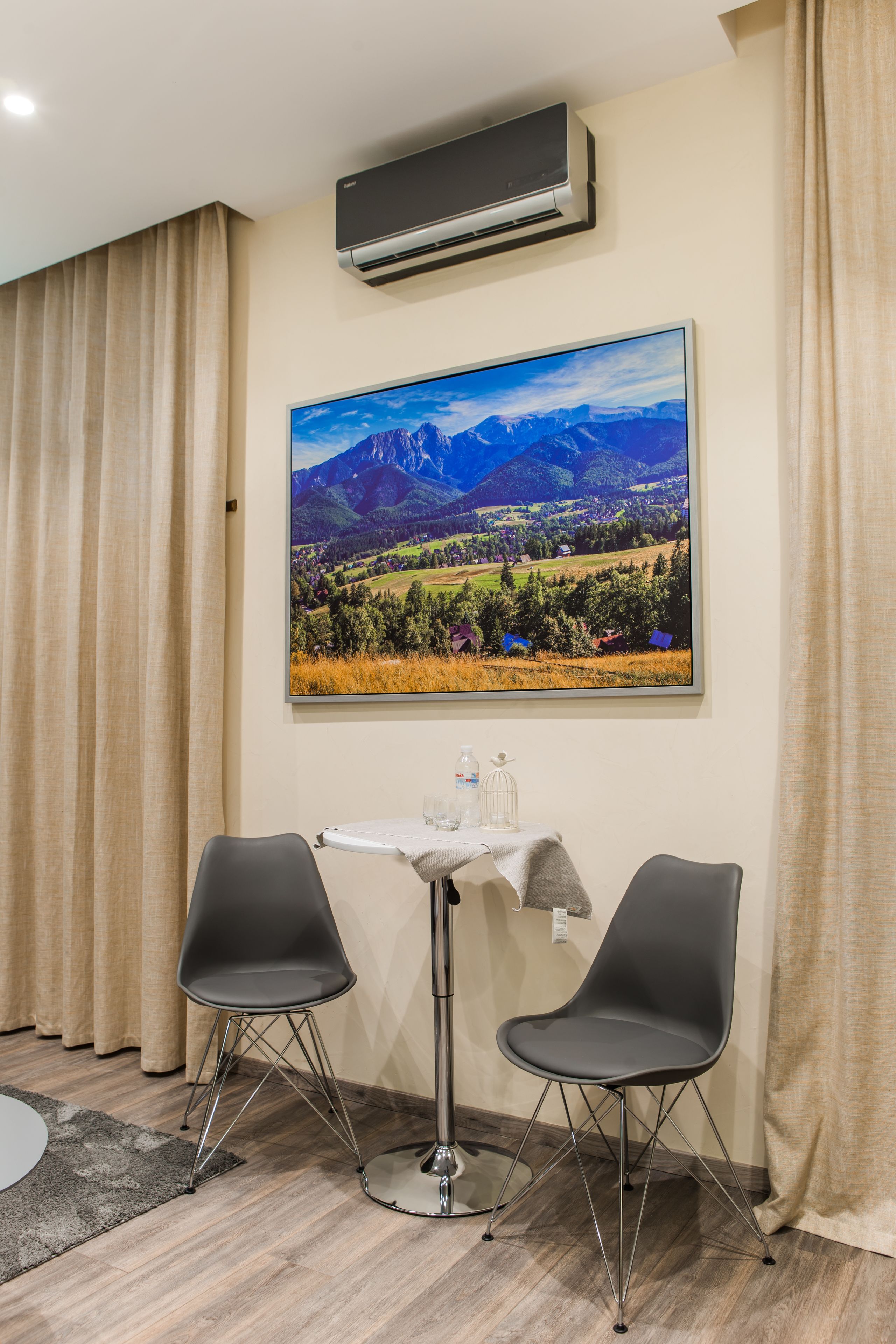Une chambre d’hôtel avec une table, deux chaises, un climatiseur et un grand tableau représentant un paysage accroché au mur.
