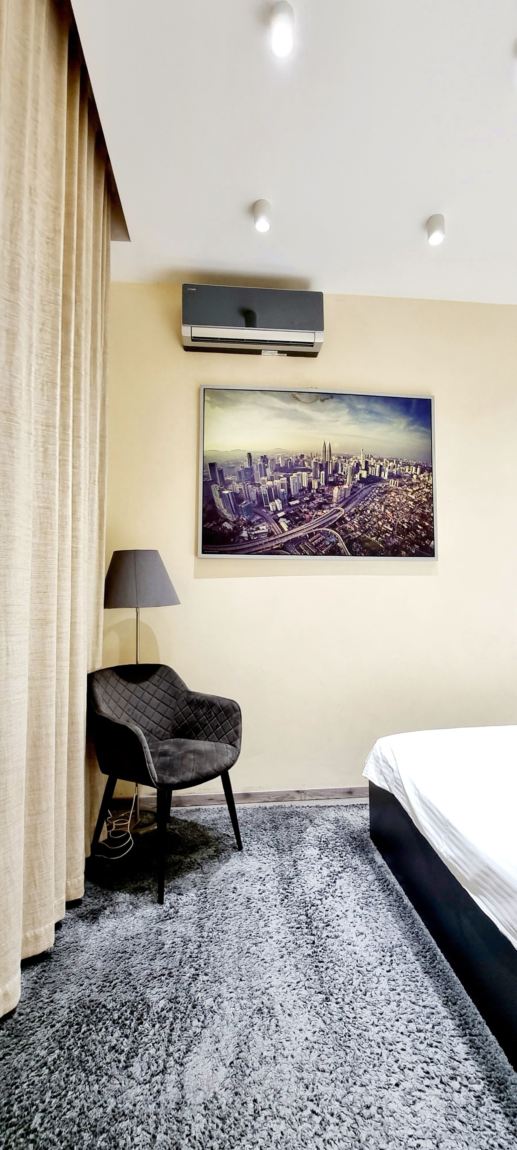 Habitación de hotel con una cama, un cuadro de una vista urbana en la pared, una silla, una lámpara y un equipo de aire acondicionado.