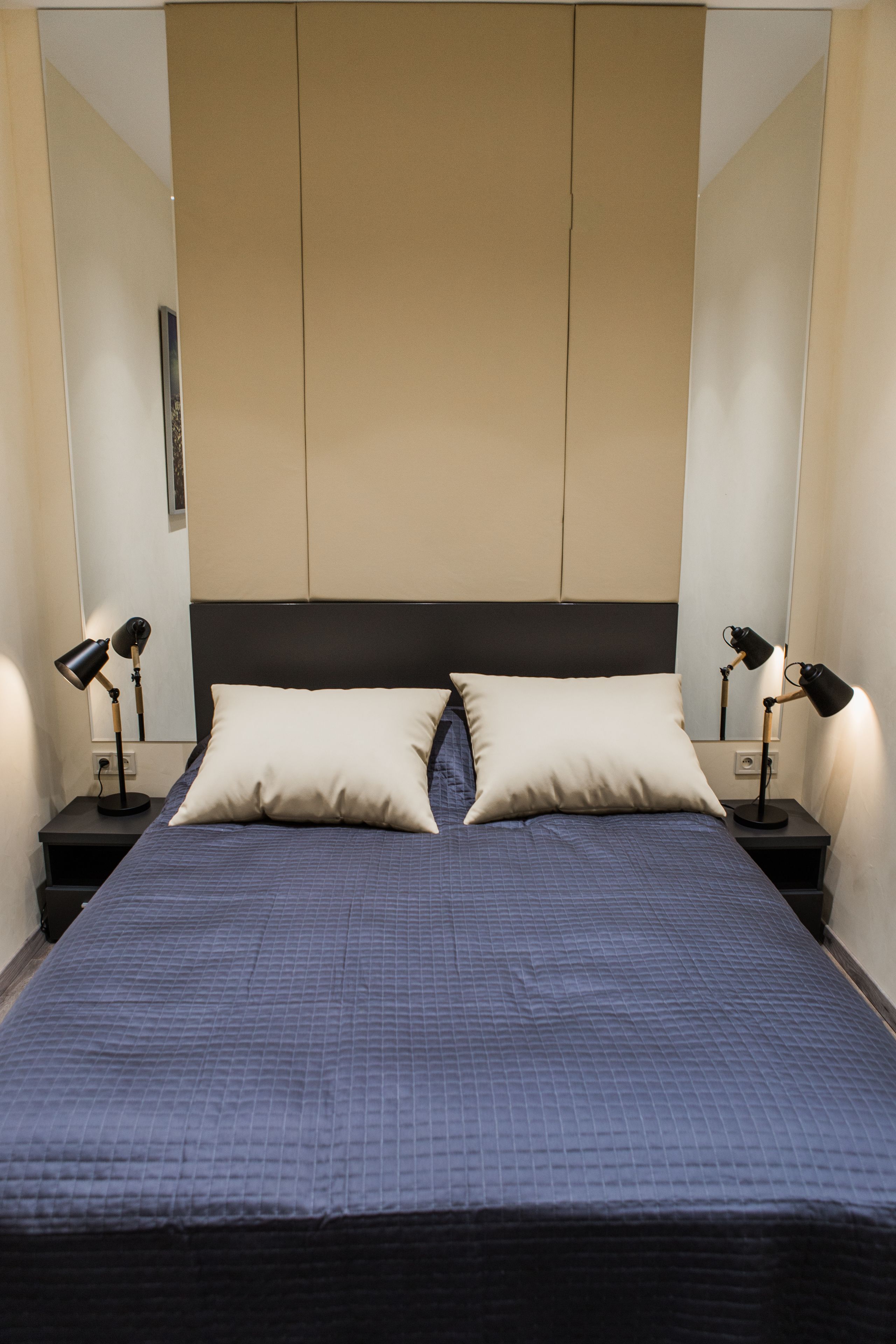 Un letto ben rifatto con una coperta blu, due lampade ai comodini e un grande specchio a parete.