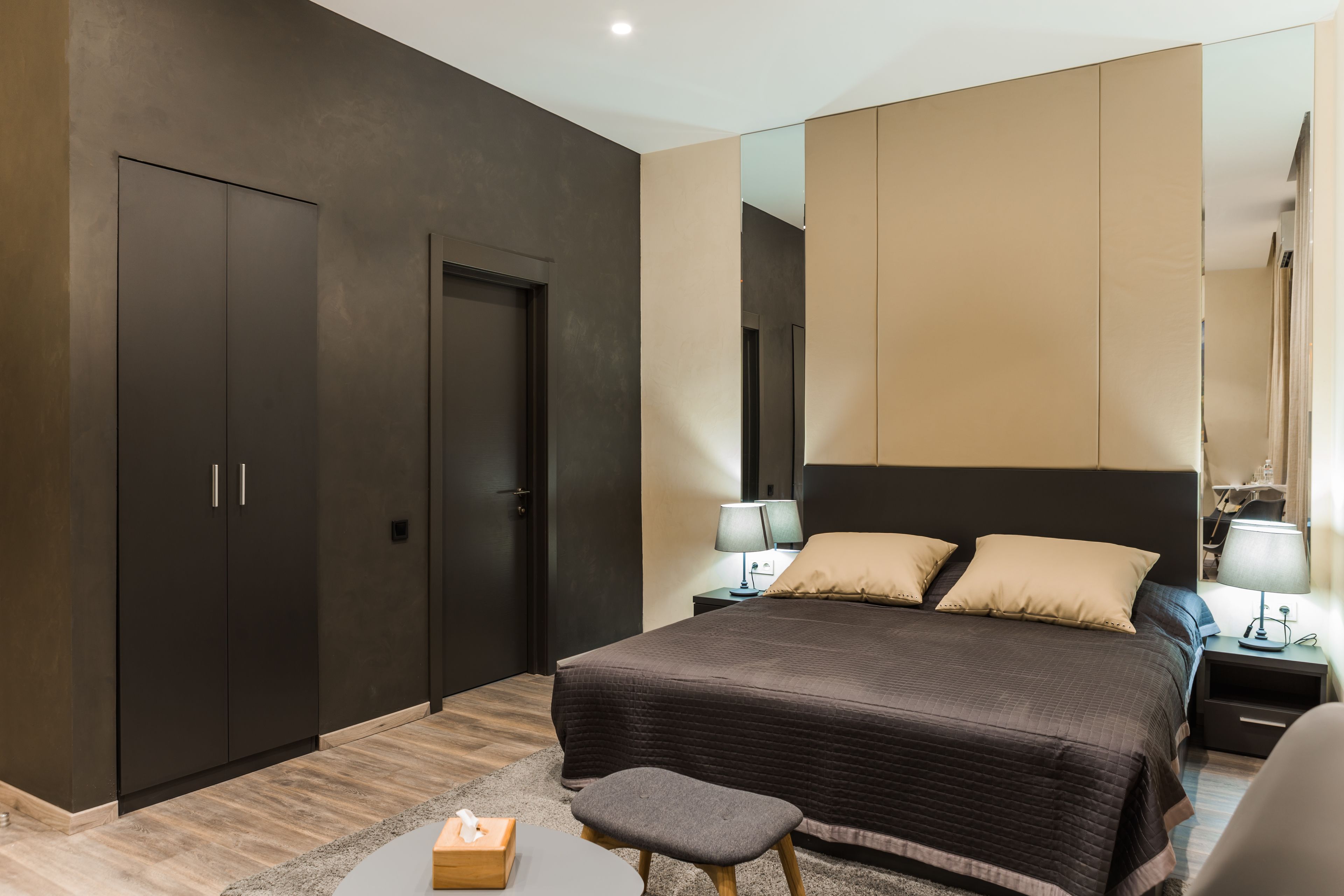 A modern bedroom with a large bed, dark wardrobe, bedside tables with lamps, and a small stool.