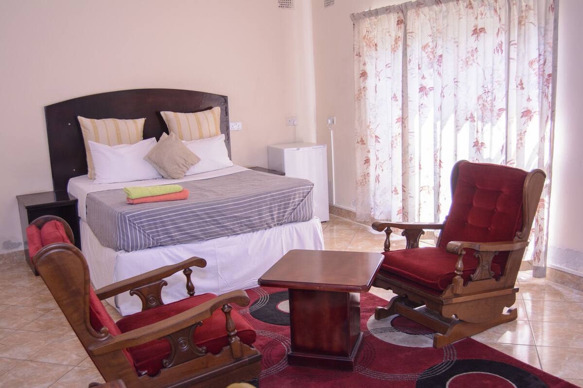 Um quarto com uma cama, uma mesa de cabeceira de madeira, uma poltrona vermelha, uma cadeira de balanço de madeira e uma pequena mesa.