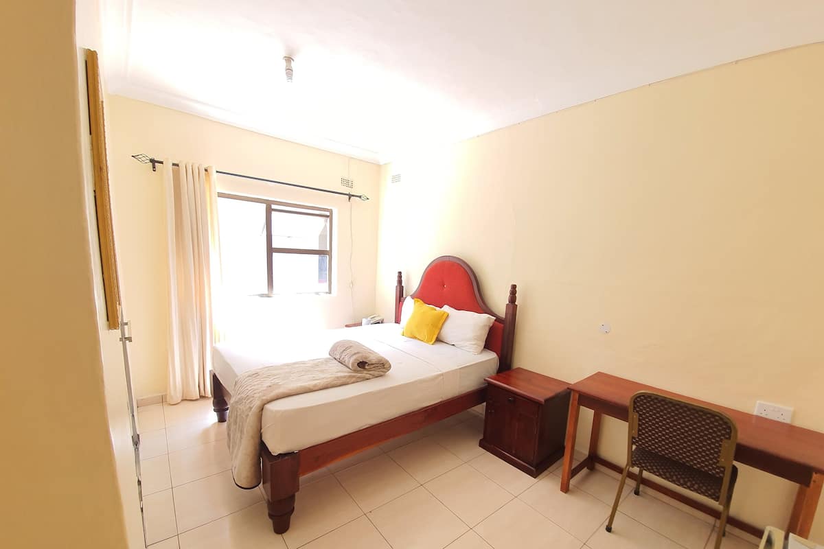 Quarto de hotel com cama, mesa de cabeceira, cadeira e janela com cortinas.