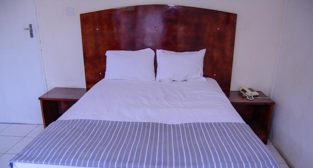 Een net opgemaakt bed met een gestreept dekbed, twee witte kussens en een houten hoofdbord. Er staan twee nachtkastjes, waarvan er één een telefoon heeft.