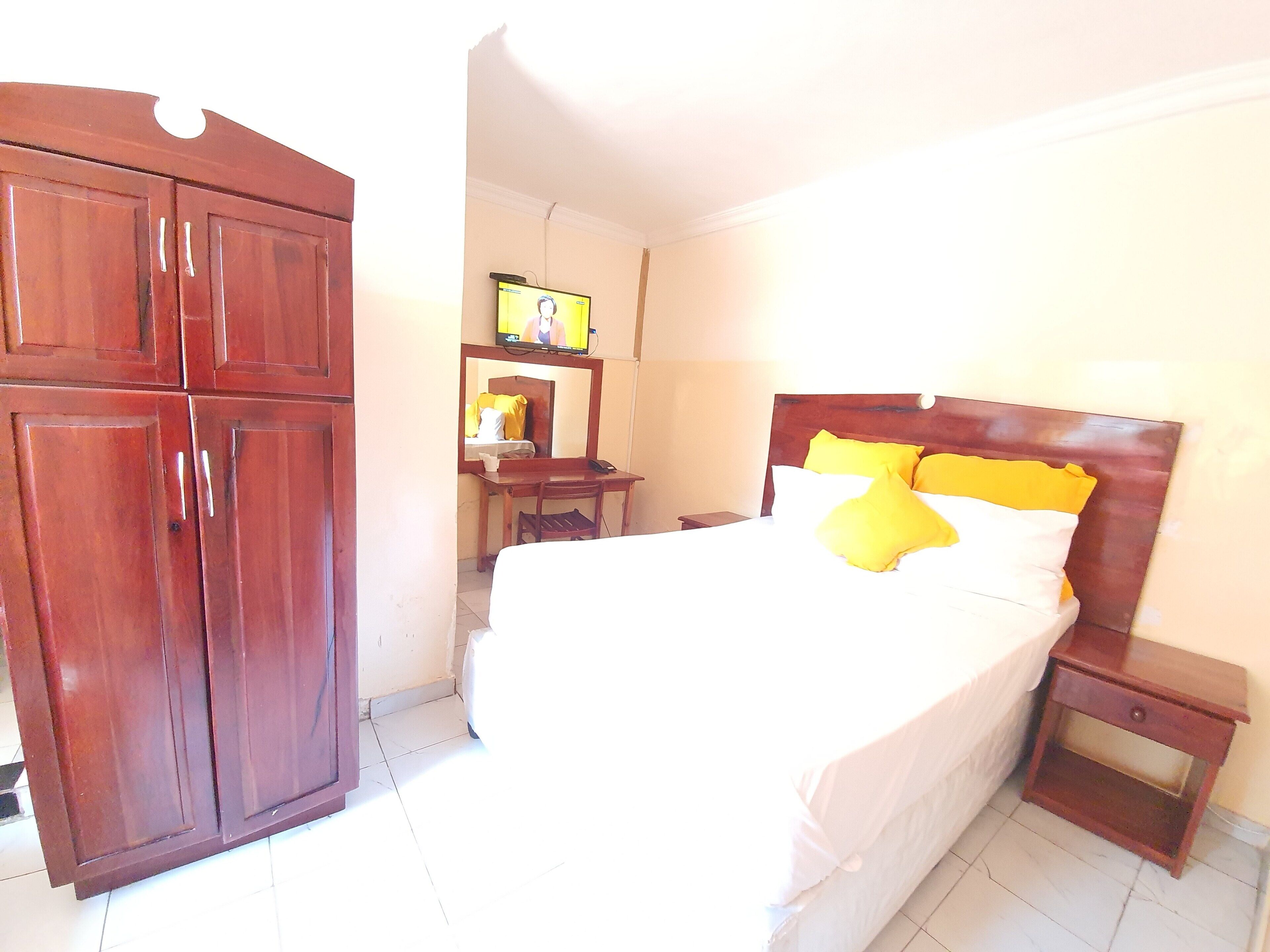 A hotel room with a bed, a wooden wardrobe, a desk with a chair, and a television on the wall.