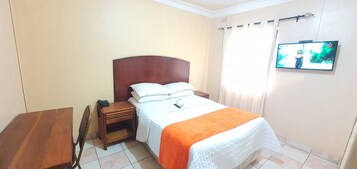 Un dormitorio con una cama, una mesita de noche, una mesa de madera y un televisor en la pared.