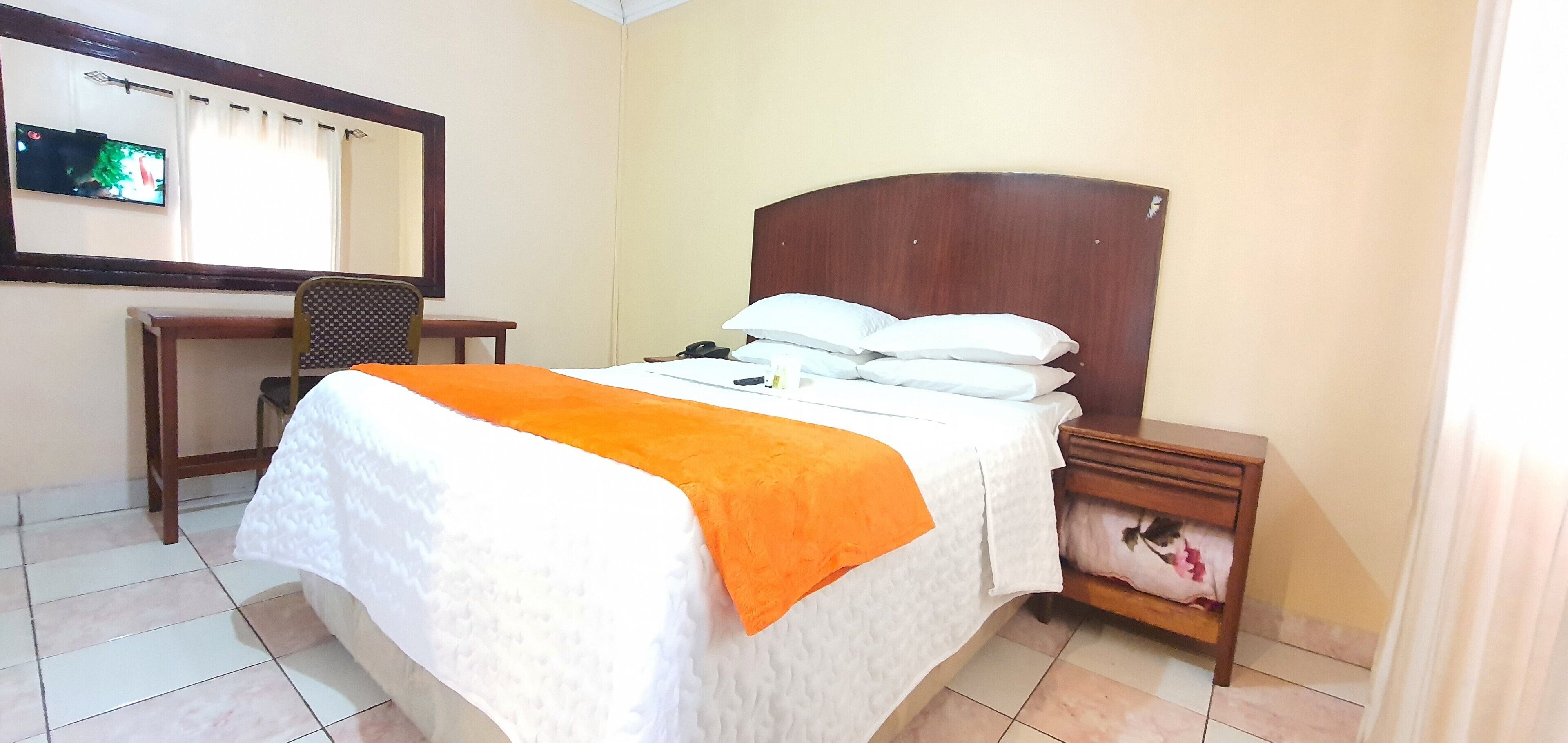 Una habitación de hotel con cama, escritorio, silla, mesita de noche y televisor en la pared.