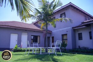 Un edificio de dos pisos con techo de tejas, una palmera y un pequeño patio con sillas.
