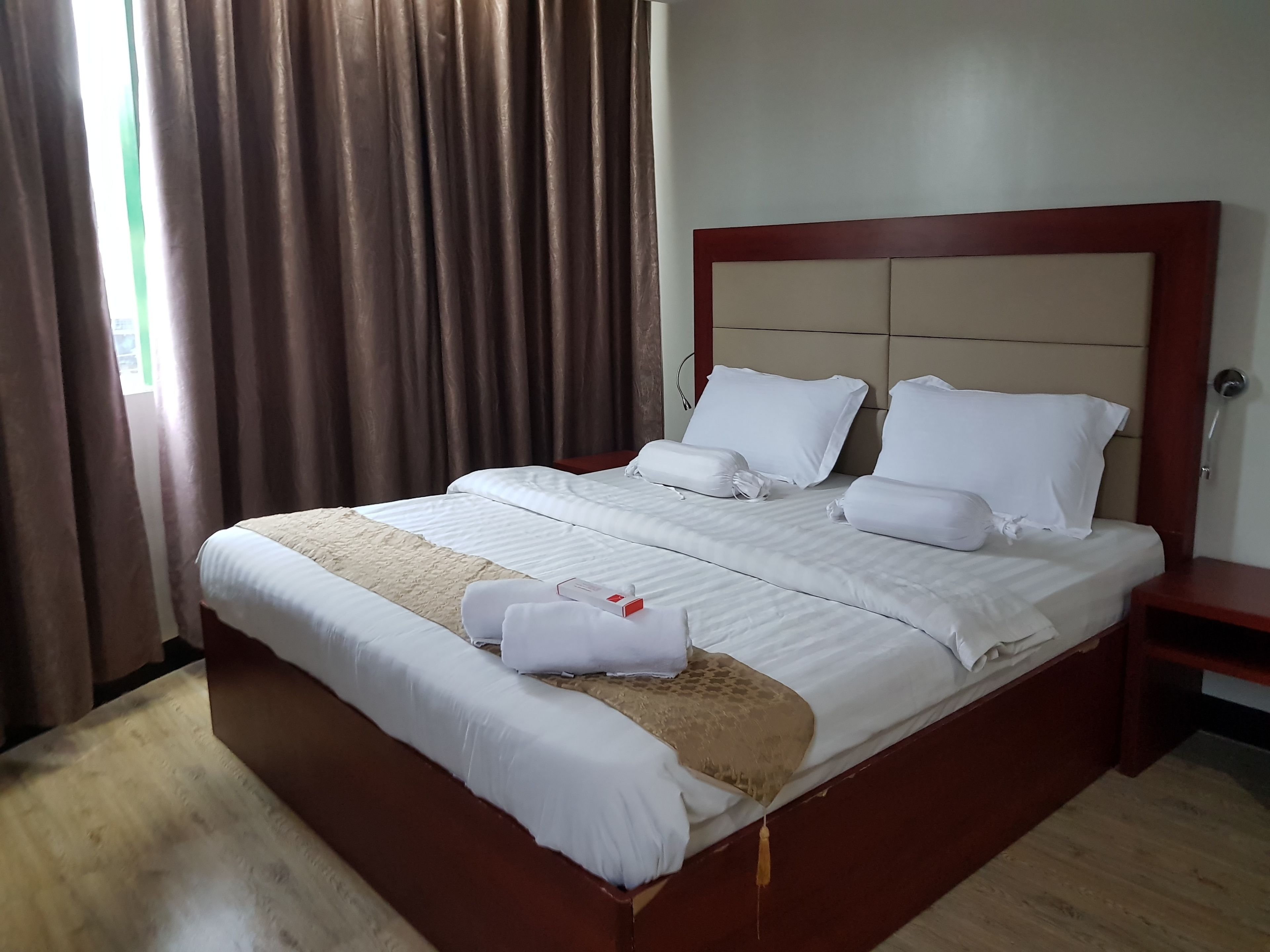 Una cama bien hecha con sábanas y almohadas blancas, un cabecero de madera y una mesita de noche de madera con un detalle rojo.