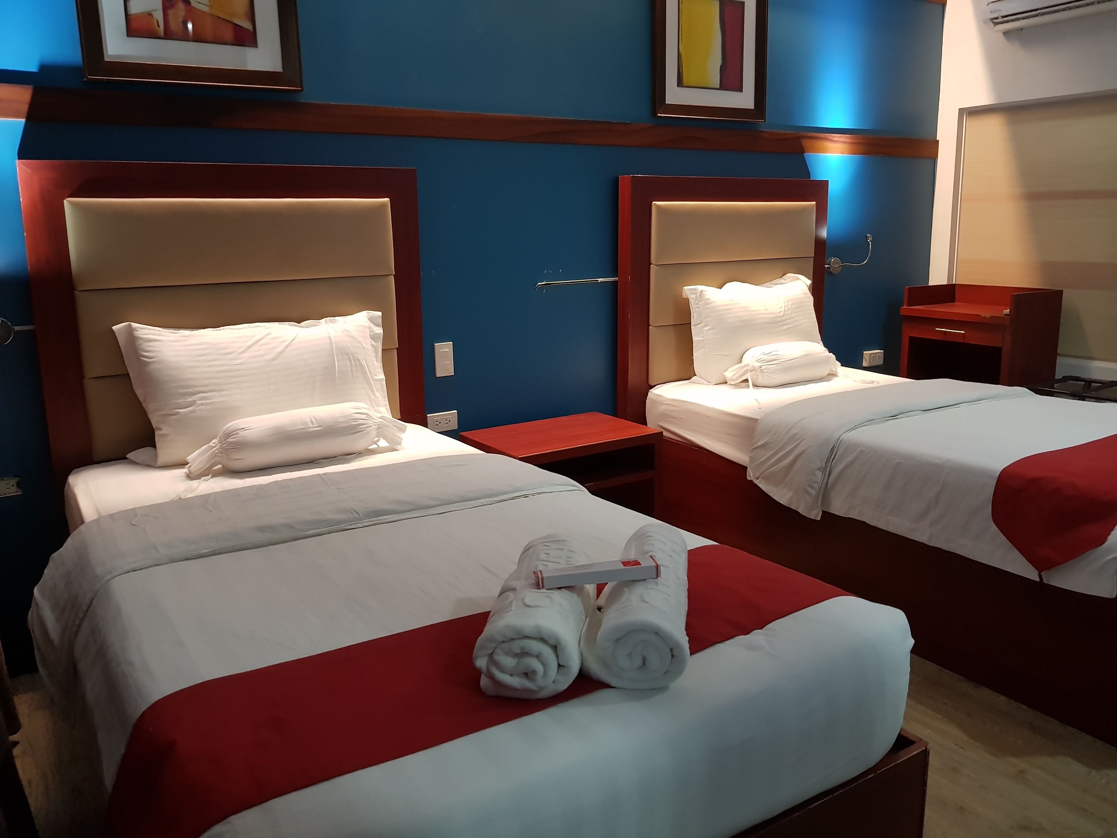 A hotel room with two beds, each with white linens and red accents. There are two nightstands, a wooden dresser, and a framed picture on the wall.