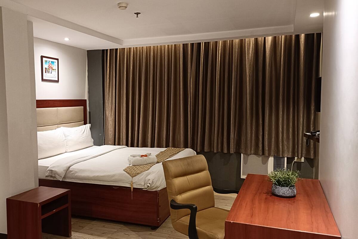 A hotel room with a bed, a desk, a chair, a small table, a potted plant, and a picture on the wall.