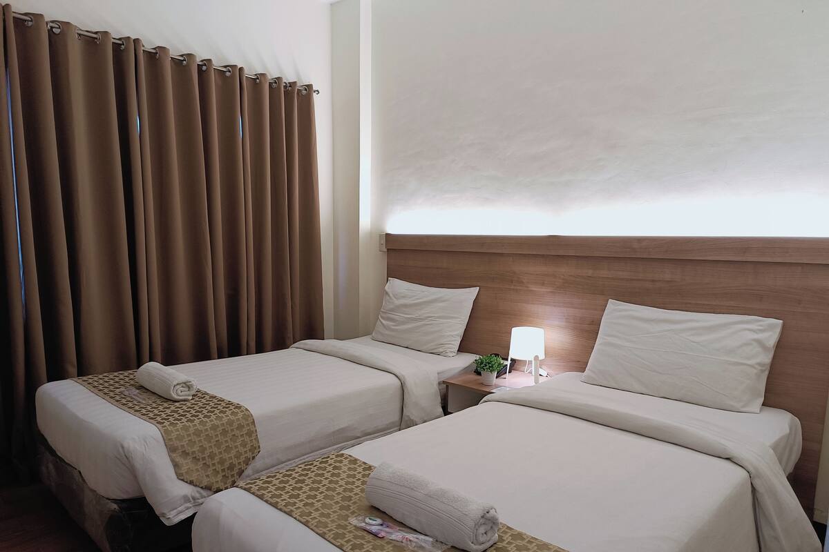 A hotel room with two beds, a wooden headboard, a nightstand with a lamp, and a window with curtains.