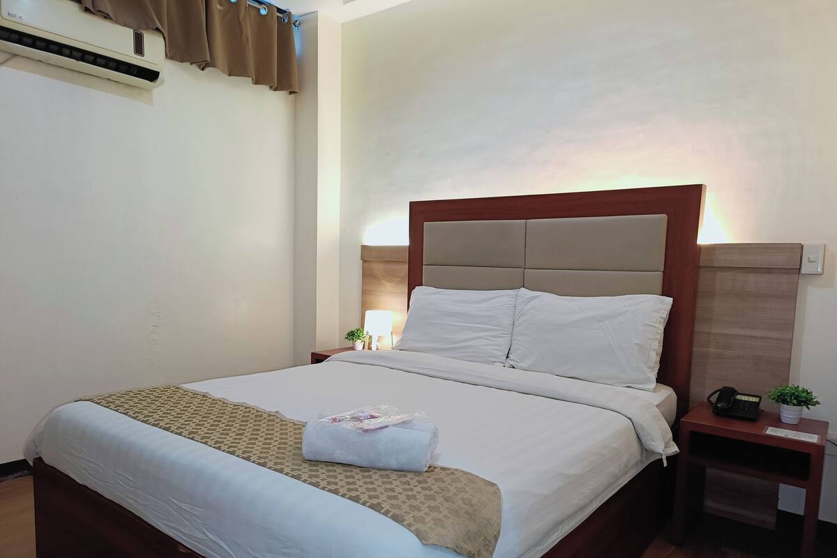 A neatly made bed with a beige headboard, a bedside lamp, and a small table with a phone and a plant.