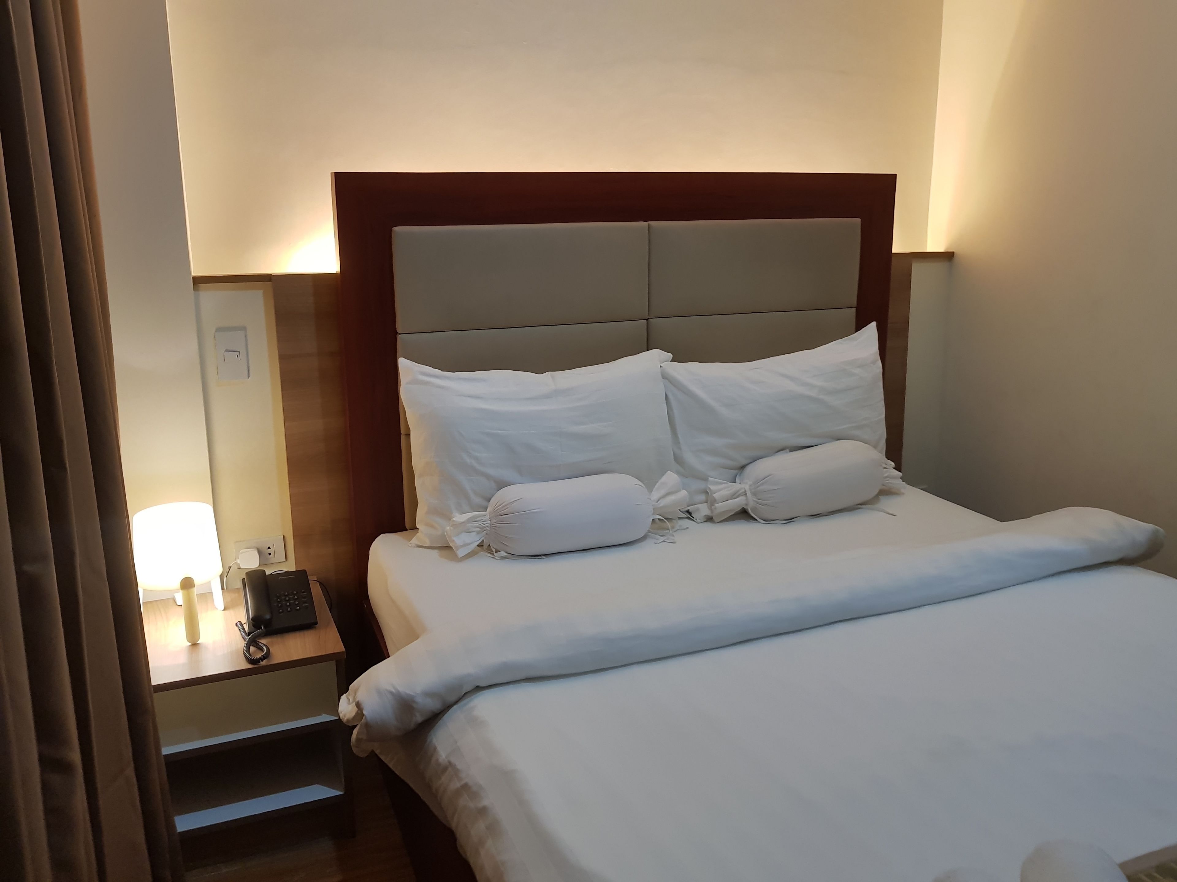 Una cama bien tendida con sábanas y almohadas blancas, una mesita de noche con lámpara y teléfono.