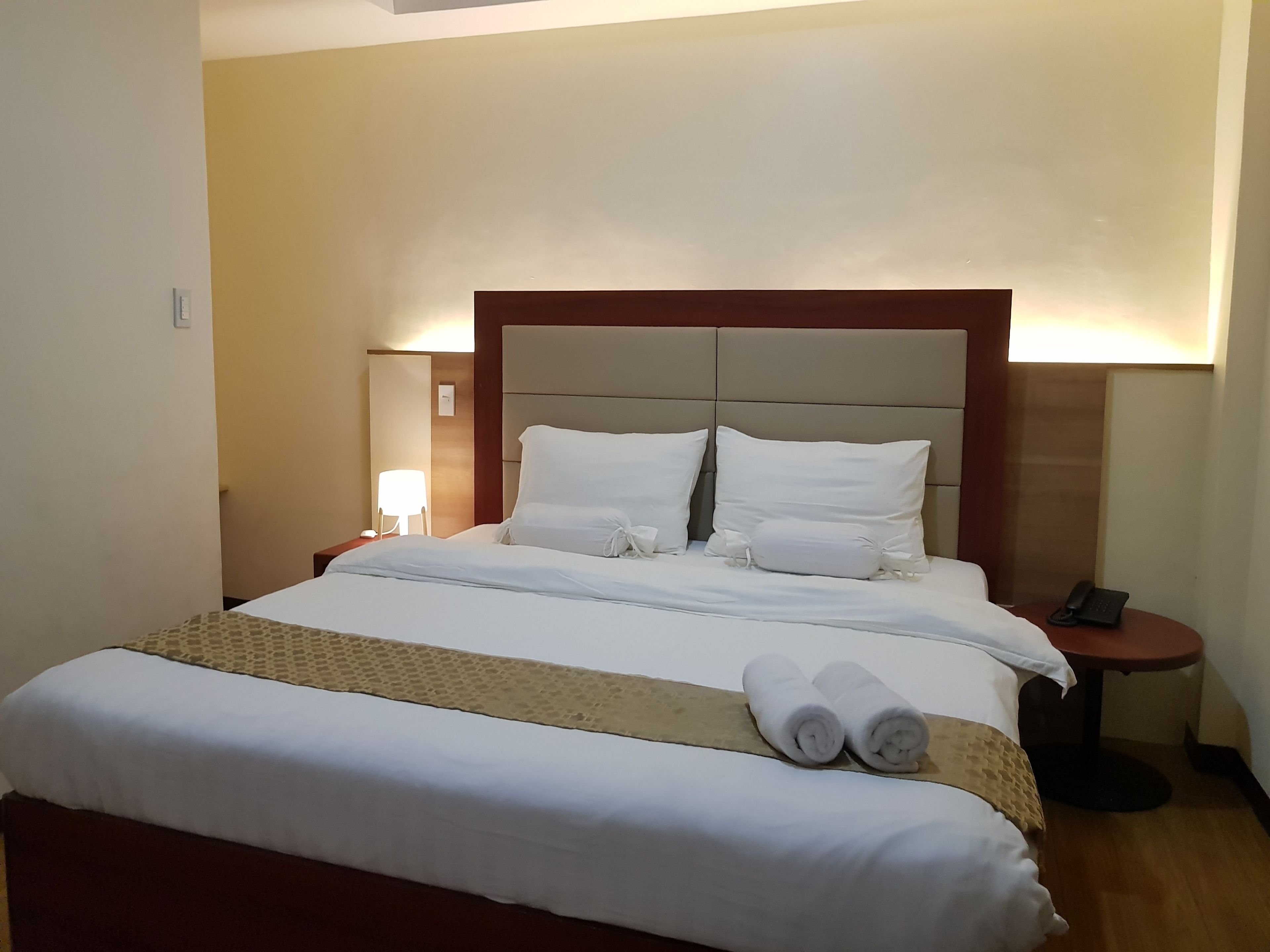 A neatly made bed with white linens and a brown headboard, a bedside lamp, and a phone on a small table beside the bed.