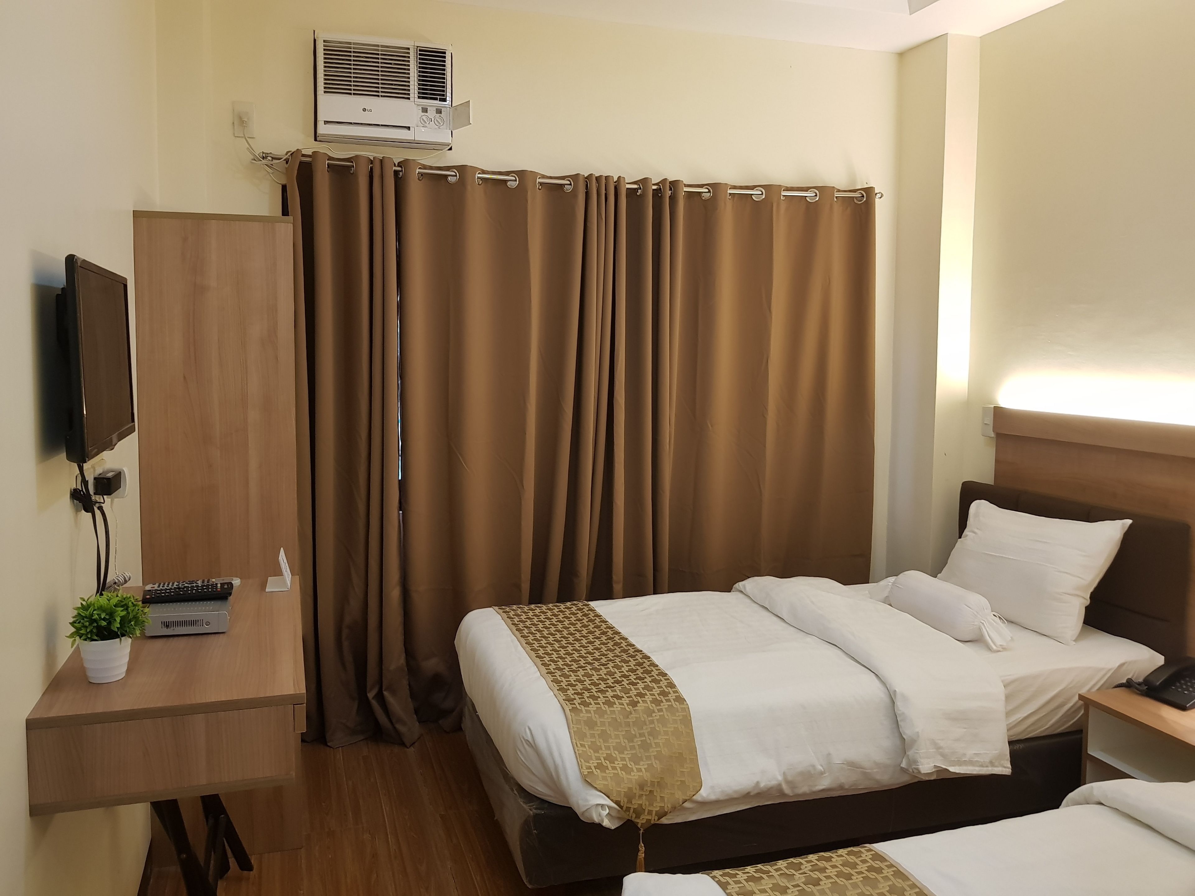 A hotel room with a bed, bedside table, TV, and an air conditioning unit.