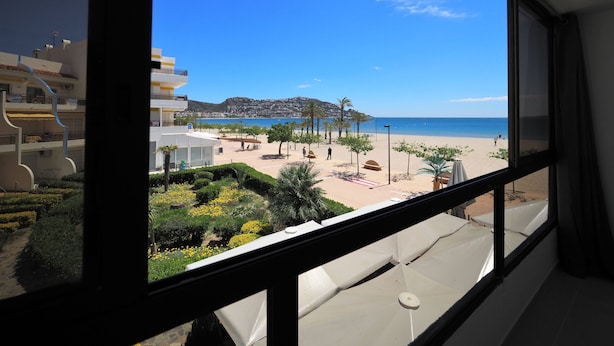 A view from a window overlooking a beach with a clear sky, palm trees, and a building with balconies.