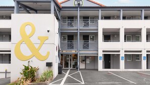 Un hotel con un grande cartello giallo a forma di '&', diversi balconi e una insegna con la scritta 'ACCUEIL' che indica la reception.
