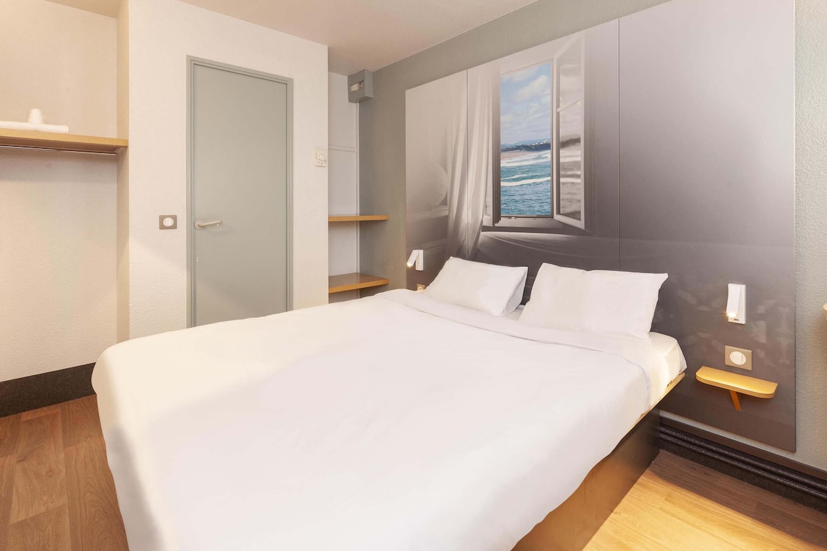 A neatly made bed with white linens, a bedside table with a lamp, and a window with a view of the sea.